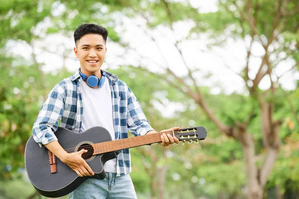 アジア系の若者が屋外でギターを弾きながらカメラで微笑む姿 — ストック写真