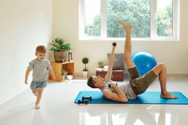 Genç baba egzersiz paspası üzerinde yerde yatıyor ve oğlu odada koşarken halterlerle egzersiz yapıyor.