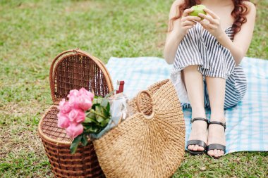 Piknik sepetinin yanındaki parkta battaniyeye oturmuş yeşil elma yiyen genç bir kadının kırpılmış görüntüsü.