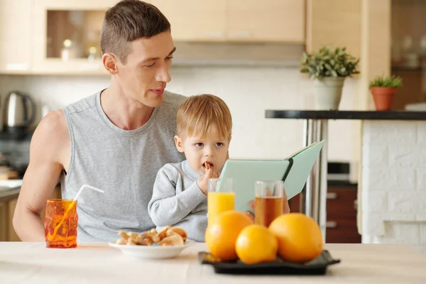 彼の小さな息子は膝の上に座って キッチンで朝食を持っている間若いお父さんはオンライン本を読んで — ストック写真