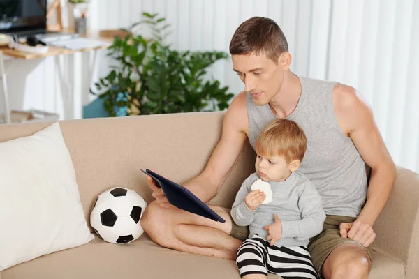 Young father sitting on sofa with his little son and watching cartoons together on digital tablet