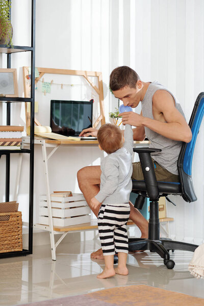 Молодой отец играет со своим ребенком во время его онлайн-работы на компьютере в комнате дома