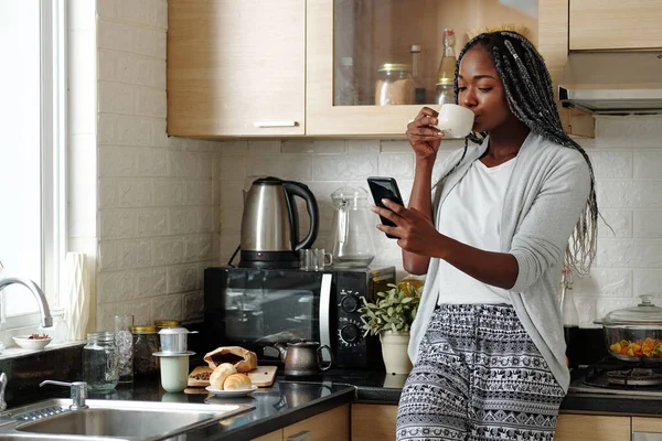 Pretty young Black woman standing at kitchen counter, drinking morning coffee and reading text messages or news on social media