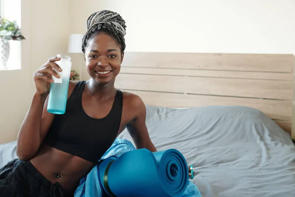 穿着辫子躺在床上 在家锻炼后喝维生素水的美丽年轻黑人妇女的画像 — 图库照片