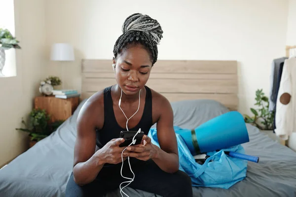非常健康的年轻黑人妇女坐在床上 用智能手机选择音乐进行训练 — 图库照片