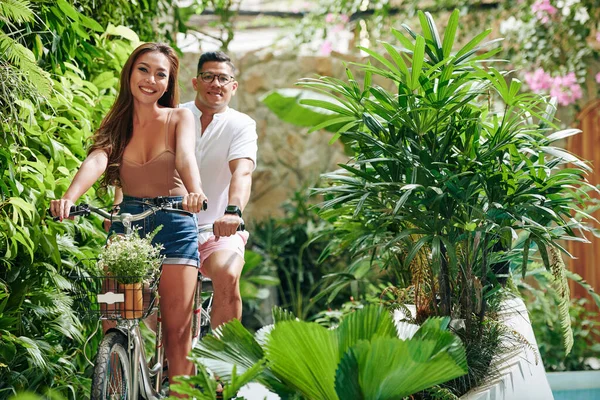 Beautiful smiling young woman in camel top and denim shorts riding tandem bicycle with her boyfriend in city park with lush plants