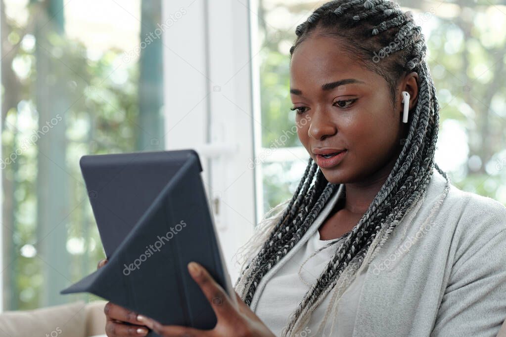 Portrait of pretty young Black woman with braided hair wearing earbuds and video calling her friend of family member