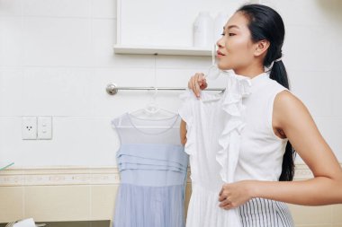 Güzel genç Asyalı kadın günün kıyafetini seçiyor ve çeşitli elbiseler deniyor.