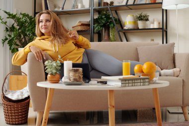 Çok güzel, genç ve iri yarı bir kadın evdeki kanepede dinleniyor, dijital tablette televizyon izliyor ve portakal suyu içiyor.