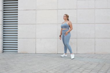 Açık havada duran gri spor sutyenli ve taytlı genç bir kadının tam boy portresi.