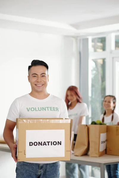 寄付された食料品の大きな段ボール箱を保持するハンサムなベトナムの若いボランティアを笑顔 — ストック写真