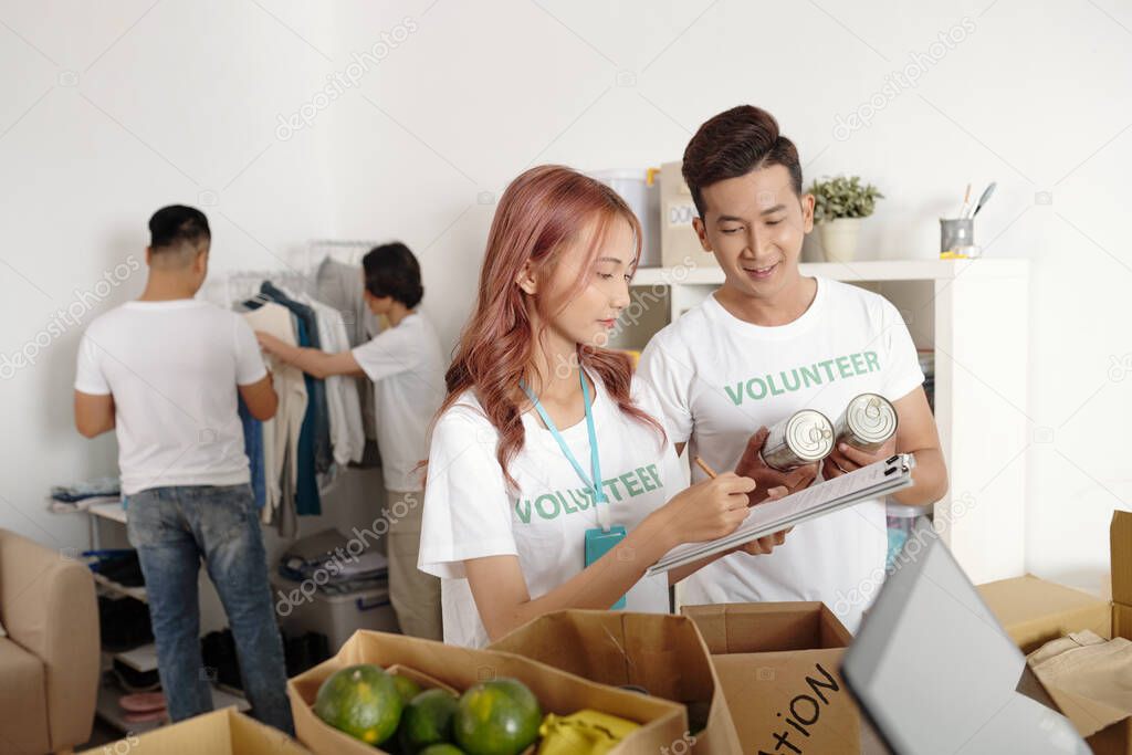 Volunteers working in charitable foundation taking notes in document and describing all food and clothes they packing for people in need