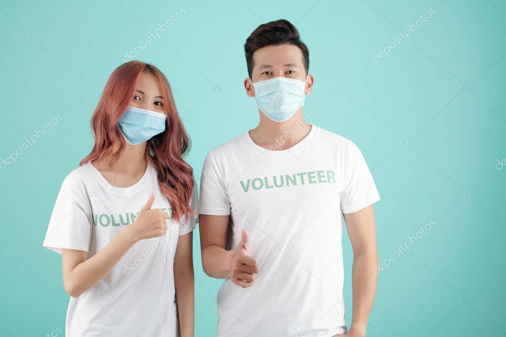 Young Asian students in volunteer t-shirts and medical masks showing thumbs-up and looking at camera