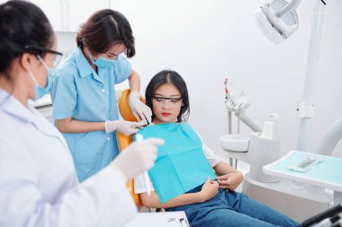 Hemşire, dişçinin tıbbi geçmişini kontrol ederken kadın hastanın göğsüne tek kullanımlık koruyucu çarşaf koyuyor.