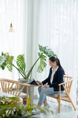 Kafe masasında oturan, kahve içen ve tablet bilgisayarda uygulama kullanan genç bir iş kadını.