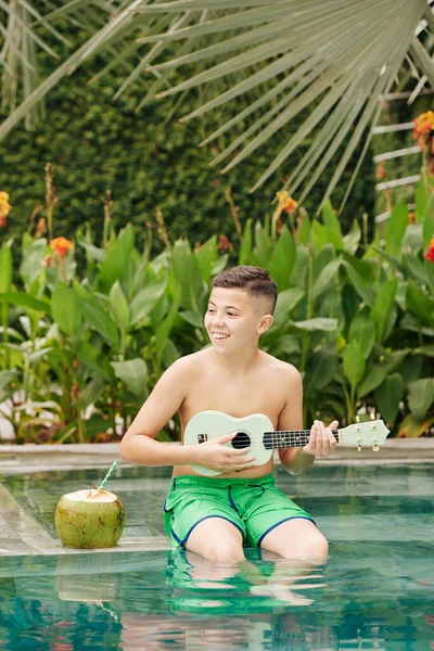 幸せな興奮した少年はプールの端に座って ココナッツカクテルを飲み ウクレレで遊ぶ — ストック写真
