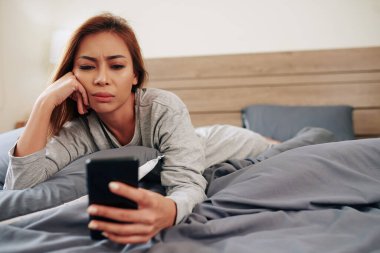 Üzgün duygusal genç kadın yatakta yatarken akıllı telefondan kötü haberler okuyor.