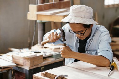 Ahşap tahtayla çalışırken keski ve çekiç kullanan konsantre Asyalı genç adam.