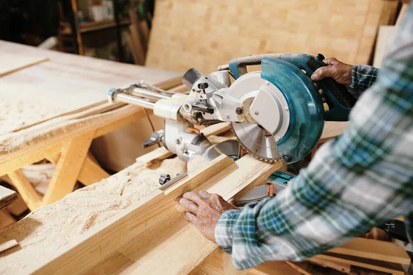 Senior carpenter cutting wood plank with circular saw when preparing details of furniture item