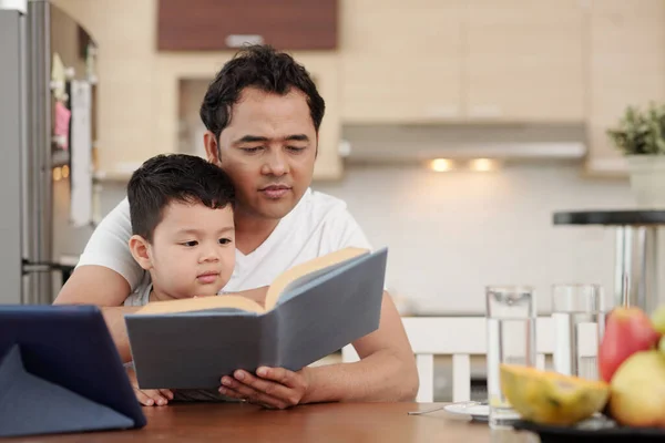 彼らは朝食後にキッチンテーブルに座っているとき 彼の小さな息子のための本を読んで気に父 — ストック写真