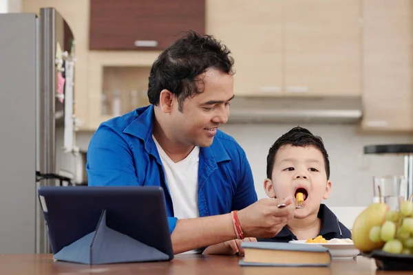 幸せなインド人男給餌彼の息子でマンゴーとバナナスライスで彼ら座っているときテーブルにデジタルタブレット — ストック写真