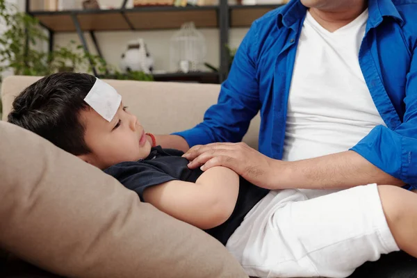 冷たいパッチがソファに横たわっている病気の子供 父は近くに座って 貧しい男の子を慰める — ストック写真