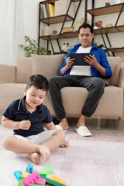 懒惰的父亲在平板电脑上看视频时 他的儿子在他前面的地板上玩彩色玩具块 — 图库照片