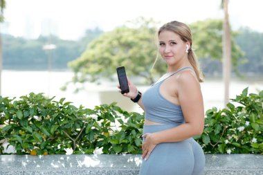 Gülümseyen genç ve kilolu bir kadın antrenmandan sonra akıllı telefondan spor salonuna bakıyor, geri dönüyor ve kameraya bakıyor.