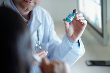 Doktor, mavi sıvı şişeyle yapılan toplantıda meslektaşına yeni bir aşı yapmanın avantajlarını anlatıyor.