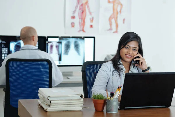 彼女の同僚は背景に胸部X線を分析するとき 若いインドの一般開業医は 電話で話をし ノートパソコンで作業笑顔 — ストック写真