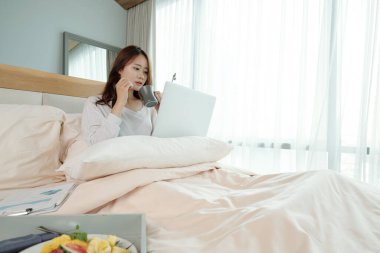 Genç Asyalı kız öğrenci yatakta çay ya da kahve içiyor ve dizüstü bilgisayardan ilginç makaleler okuyor.