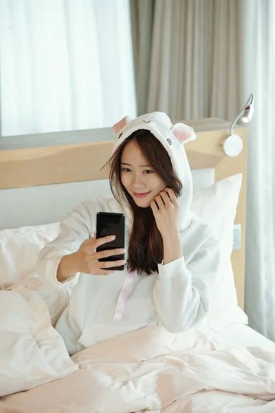 穿着睡衣坐在床上 在社交媒体上自言自语的漂亮微笑的年轻女人 — 图库照片