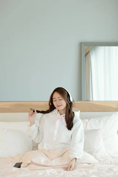 快乐而美丽的年轻亚洲女人 坐在床上 耳机里享受着美妙的音乐 — 图库照片