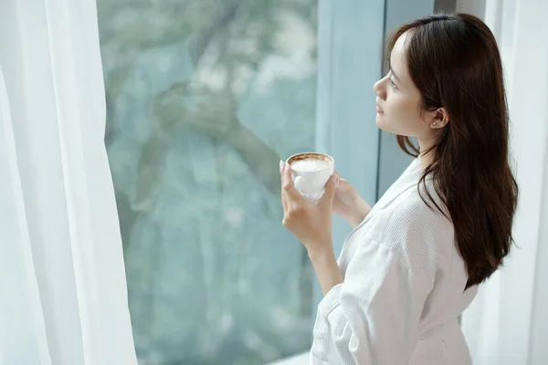 Pensiv Ung Kvinna Dricker Kopp Morgonkaffe Och Tittar Genom Lägenhetsfönstret — Stockfoto