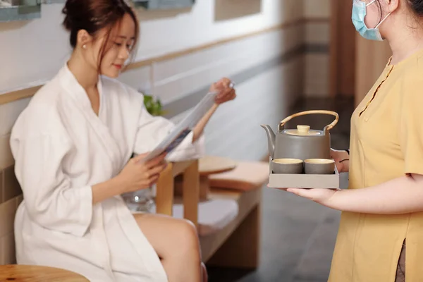 Woman reading menu in spa salon, manager in protective mask bringing her pot of herbal tea and cups