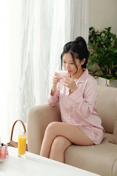 魅力的な若いです女性でシルクピンクパジャマ座っていますソファに自宅でとにおいの瓶の保湿ボディクリーム — ストック写真