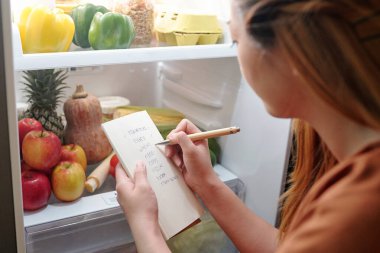 Genç bir kadın buzdolabının önünde duruyor ve alışveriş listesindeki her şeyi alıp almadığını kontrol ediyor.