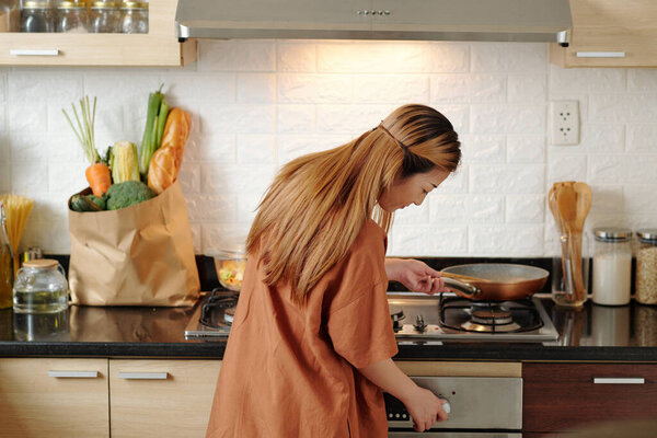 Молодая женщина включает газовую плиту, чтобы жарить овощи или сделать соус макарон на ужин, вид сзади