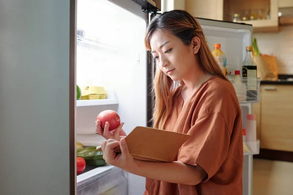 若い女性チェック彼女持っているかどうか十分な果物で冷蔵庫と作るショッピングリスト — ストック写真