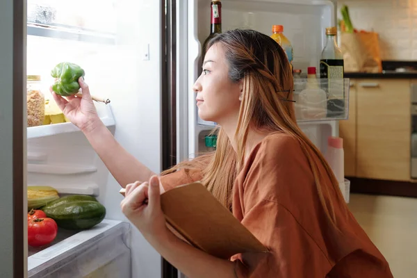 漂亮的年轻女人在列购物单的时候会检查冰箱架子上的蔬菜 — 图库照片