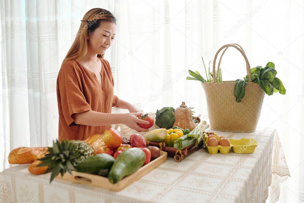 Smiling young woman unpacking bags of groceries she bought at local market and putting fruits and vegetable on trays