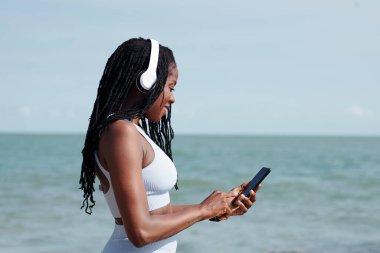 Gülümseyen, kulaklıklı genç bir kadın, akıllı telefon uygulaması üzerinden eğitimi için müzik seçiyor.