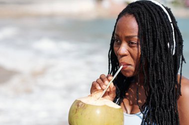 Rasta giymiş, kulaklıkla müzik dinleyen ve hindistan cevizi kokteyli içen gülümseyen genç bir kadının portresi.