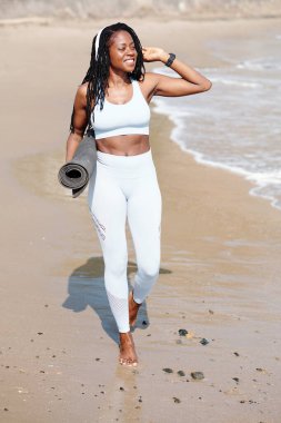 Elinde yoga minderiyle sahilde yürüyen ve deniz manzarasının keyfini çıkaran zinde, genç bir kadın.