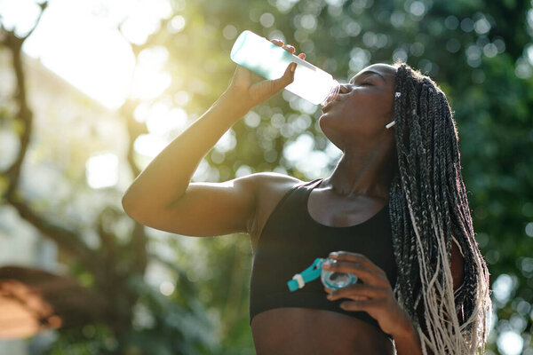 красивая молодая женщина с плетеными волосами пьет пресную воду в солнечных лучах после тренировки в парке