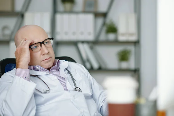 Confused mature Caucasian doctor in lab coat wearing stethoscope around neck massaging temple while analyzing research data on computer