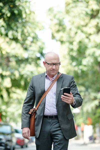 Serious busy mature bald businessman with leather bag using smartphone while working on move in summer