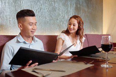 Gülümseyen genç bir çift şarap içip menü okuyorlar. Restoranda akşam yemeği için ne sipariş edeceklerine karar veriyorlar.