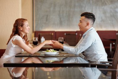 Birbirlerine aşık genç bir çift restoran masasında oturmuş, el ele tutuşuyorlar ve birbirlerine bakıyorlar.