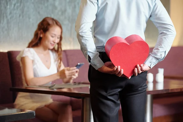 男人把礼物藏在心形盒子里 给坐在咖啡桌旁 用智能手机查看短信的女朋友看 — 图库照片